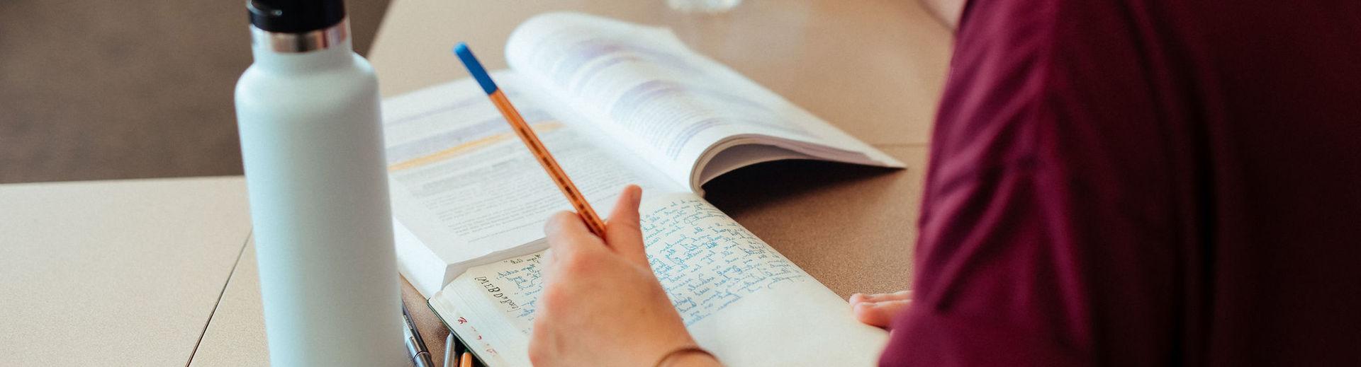 Female student is diligently taking notes while reading a highlighted text book.