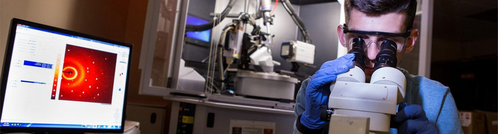 Chemistry student looking through a microscope in a lab surrounded by computer and equipment.