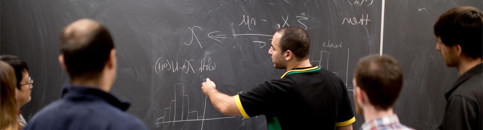 Mathematics professor writing on a chalkboard with students gathered around