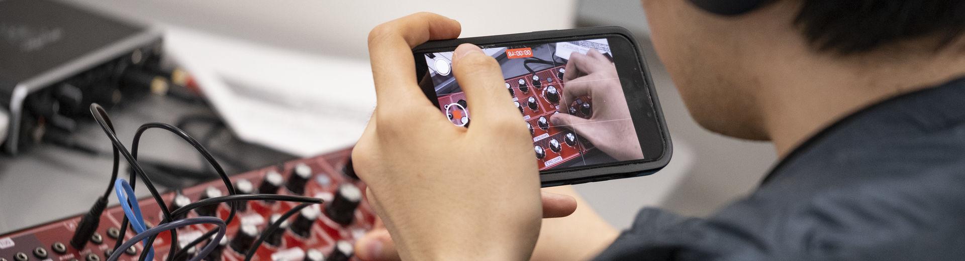 A student records a video while adjusting the dials on a synthesizer.