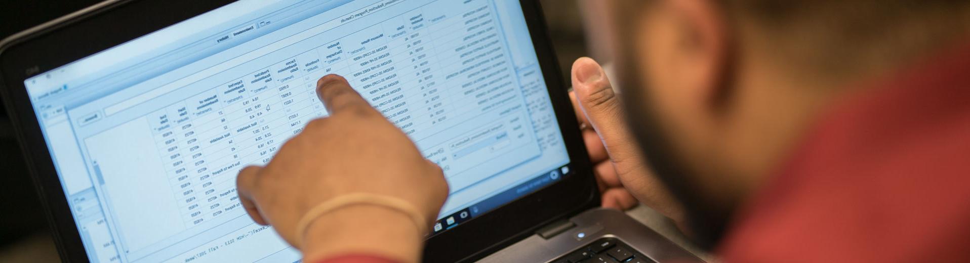 A person wearing a red shirt points to a line item on a spreadsheet displayed on a laptop screen.