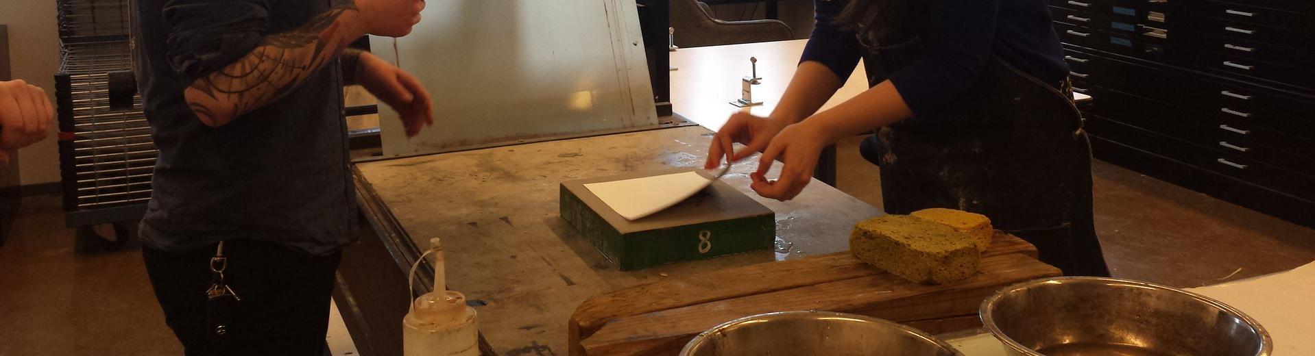 Two students look over a table where one student is peeling a sheet of paper from a block.