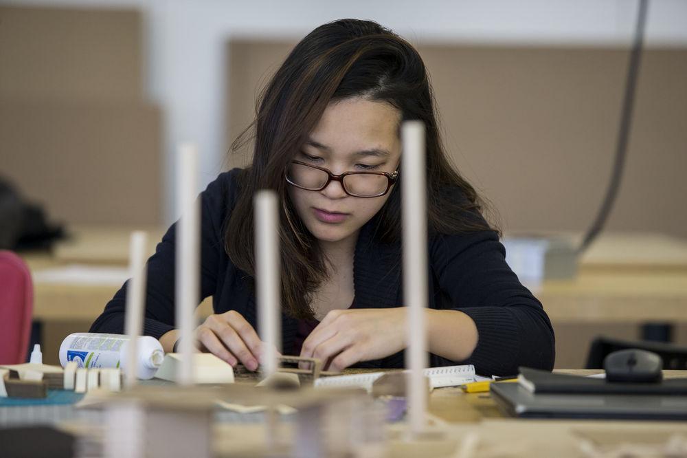 A woman with black hair and glasses works at a table with small pieces of cardboard.