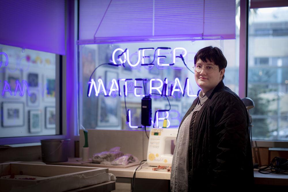 A person with glasses st和s next to a sewing machine with a backwards neon sign behind them.