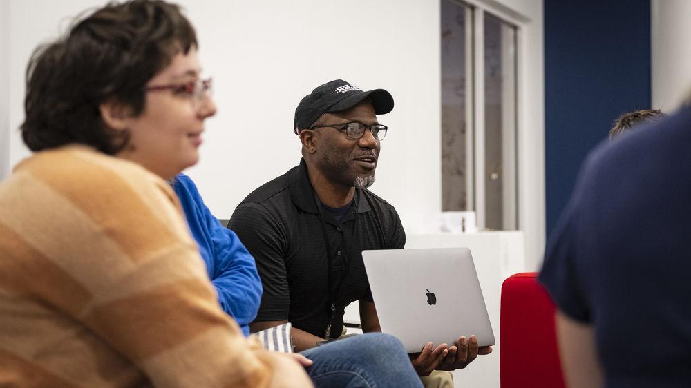 A man wearing a black baseball cap and holding his laptop sits with other students.