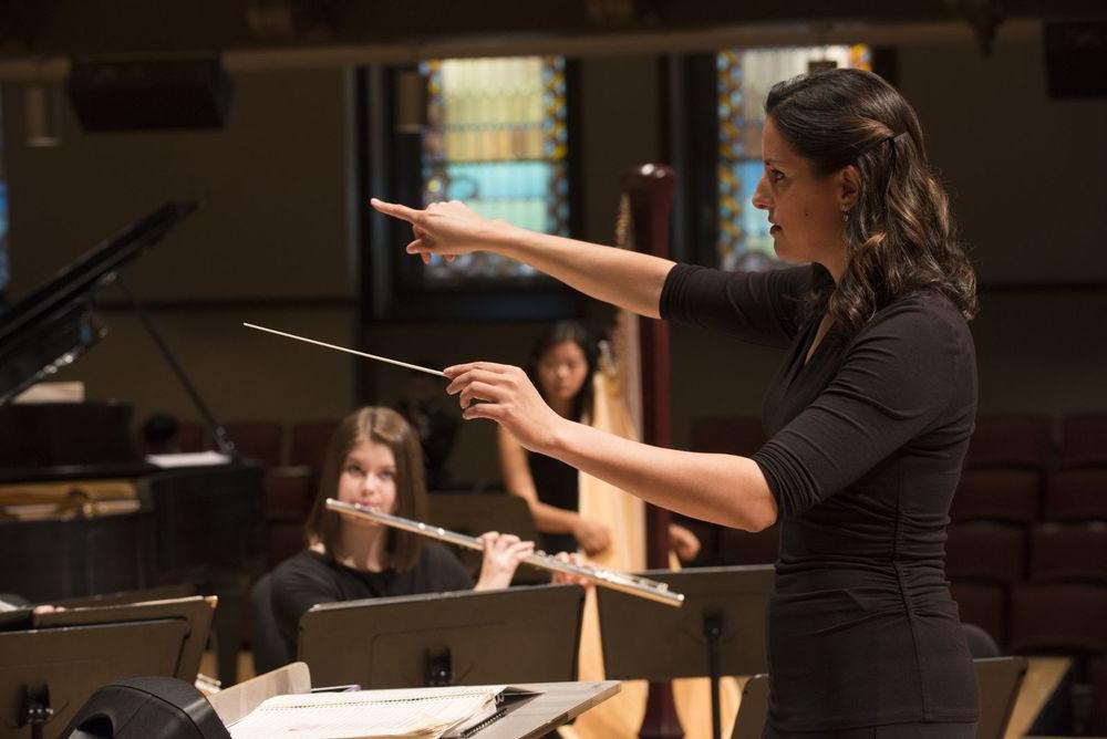 A woman conducts an orchestra with students playing flute 和 harp in the background.