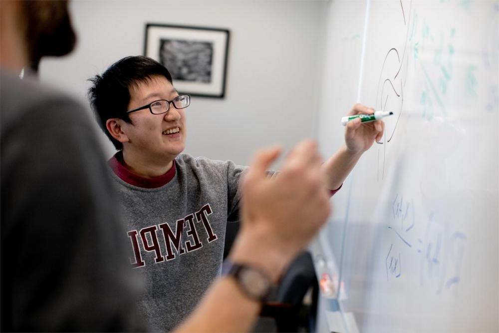 Student writing out math equation on white board with College of Science and Technology professor looking on in a Temple classroom. 